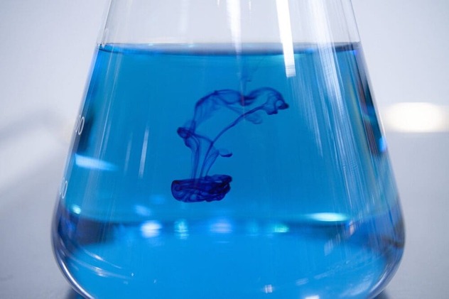 Dunkelblaue Farbtropfen verteilen sich schlierenförmig in der blauen Flüssigkeit eines Erlenmeyerkolbens